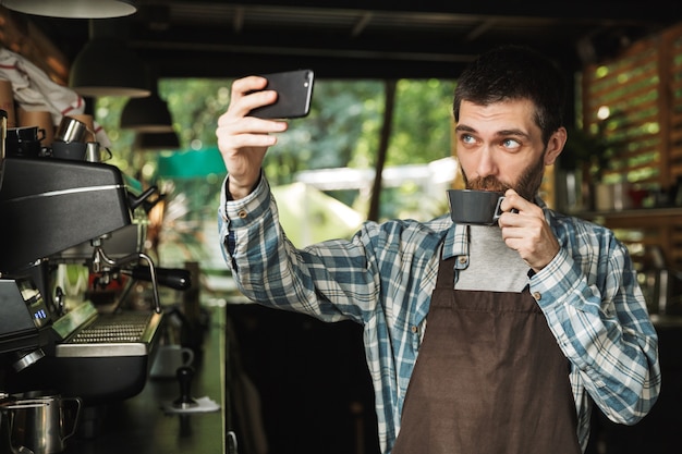 Portrait of unshaved barista man wearing apron taking selfie photo with cup of coffee while working in street cafe or coffeehouse outdoor