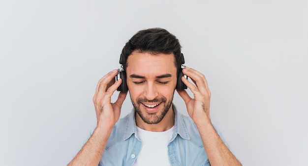 Portrait of unshaven handsome young Caucasian male with cheerful expression closes eyes as feels enjoyment and happiness holding headphones with both hands listening music in studio Copy space