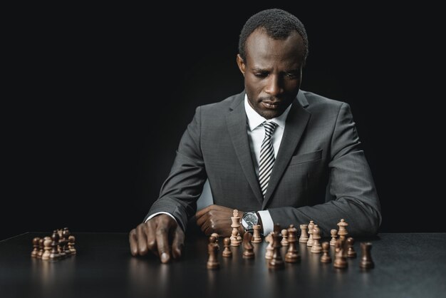 Portrait of upset african american businessman looking at chess figures isolated on black