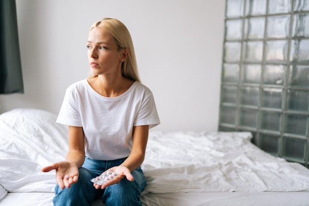 Portrait of upset blonde young woman holding pills in hands and thinking about taking drugs sitting on bed in light room at home pensive looking away