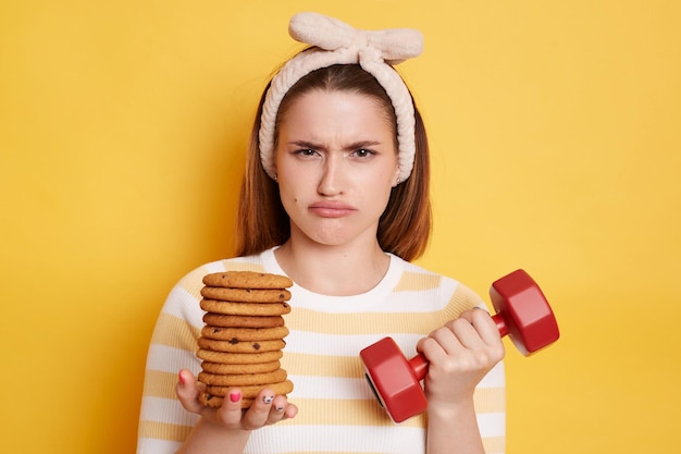 Portrait of upset sad woman with pout lips wearing striped shirt and hair band holding red dumbbell and cookies needs to choose junk food or sport posing isolated over yellow background