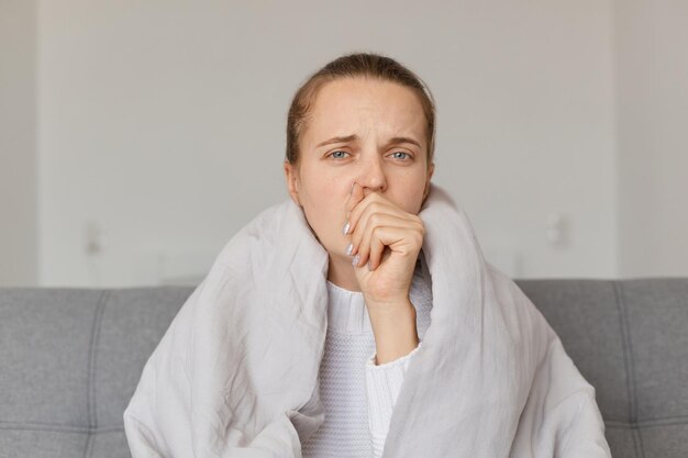 Portrait of upset sick woman with headache sitting under the blanket coughing keeping hand near mouth having sad facial expression having influenza symptoms