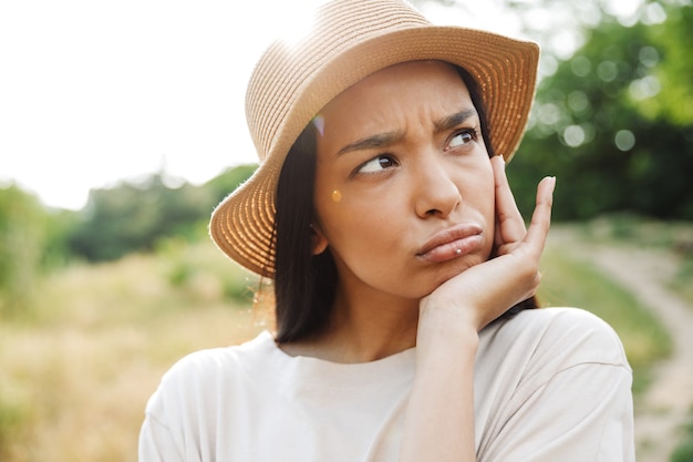 Portrait of upset woman wearing straw hat and lip piercing frowning while walking in green park