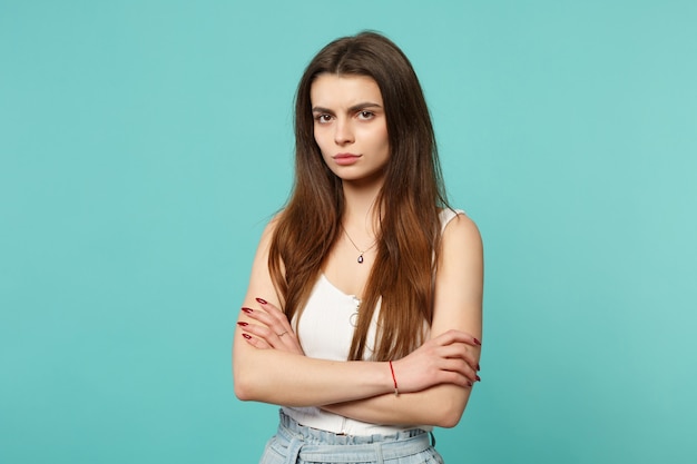 Portrait of upset young woman in light casual clothes looking camera, holding hands crossed isolated on blue turquoise wall background. People sincere emotions, lifestyle concept. Mock up copy space.