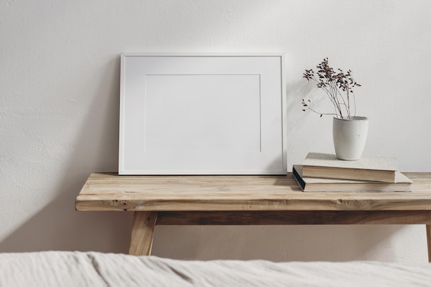 Portrait white frame mockup on vintage wooden bench table Little ceramic vase with dry hypericum plant on pile of books Blurred beige linen bed in front Modern Scandinavian interior