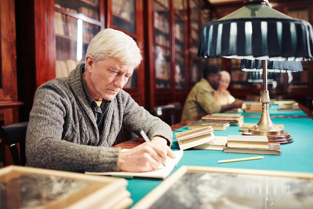 Portrait of white haired senior man studying in classic library by lamp light copy space