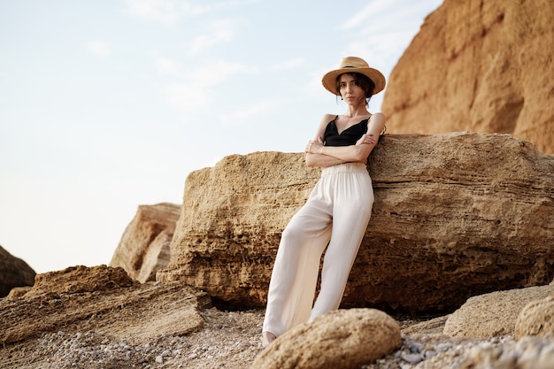 Portrait of woman in black bralette and white trousers leaning on rock at beach