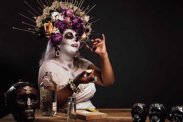 Portrait of woman in catrina makeup wearing wedding dress drinking tequila in a bar Colorful skulls on the table