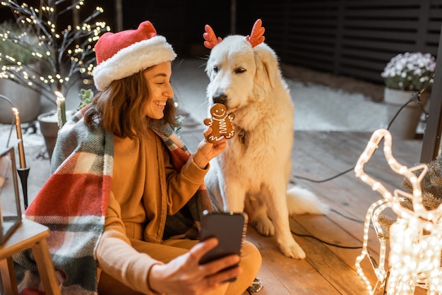 Portrait of a woman in christmas hat and plaid with her cute dog celebrating a New Year holidays on the beautifully decorated terrace at home, feeding dog with gingerbread cookies