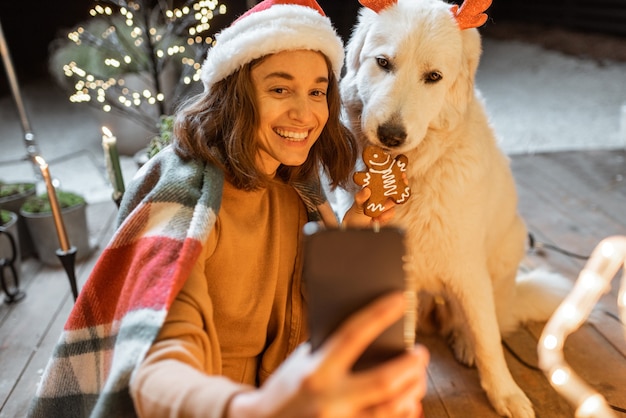 Portrait of a woman in christmas hat and plaid with her cute dog celebrating a New Year holidays at home, feeding dog with gingerbread cookies and making selfie photo