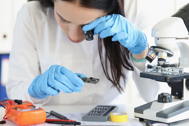 Portrait of woman examine part of digital device with magnifying glass. Female wants to see tiny details through loupe. Tech, repair service, lab concept