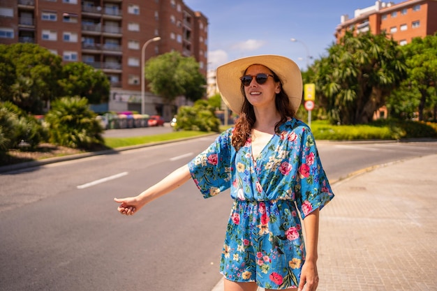 Portrait of a woman in floral summer dress and hat hitchhiking next to a street