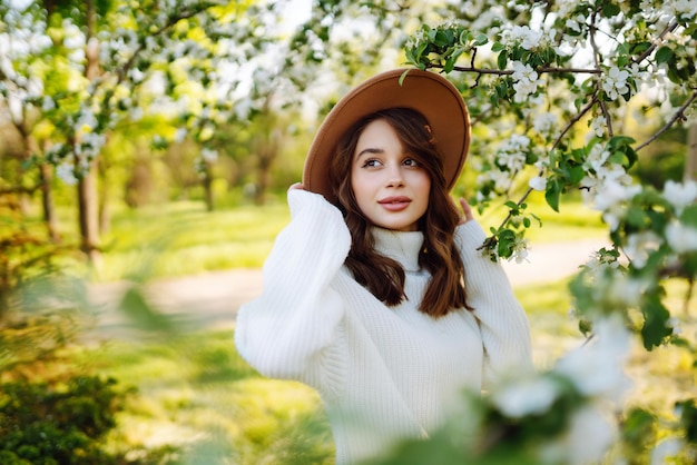 Portrait of Woman In hat posing near flowering tree Smiling young woman enjoying smell of flowers