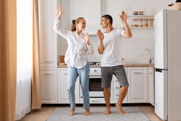 Portrait of woman and man wearing jeans and white shirts expressing happiness and dancing with raised arms looking at each other with smile posing in kitchen