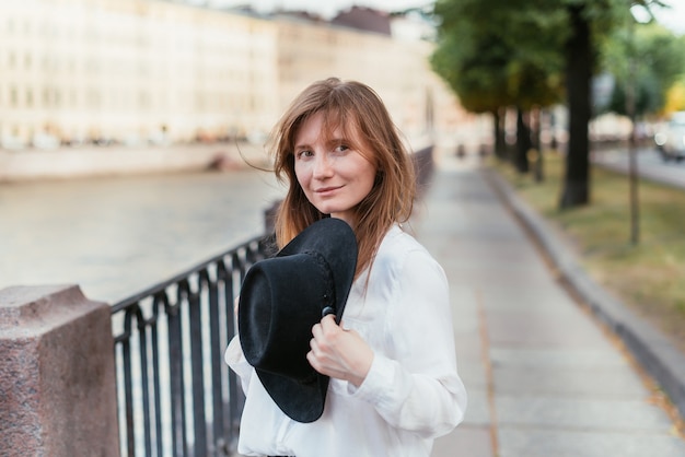 Portrait of a woman near the embankment in the city in the summer holding a hat and smiling