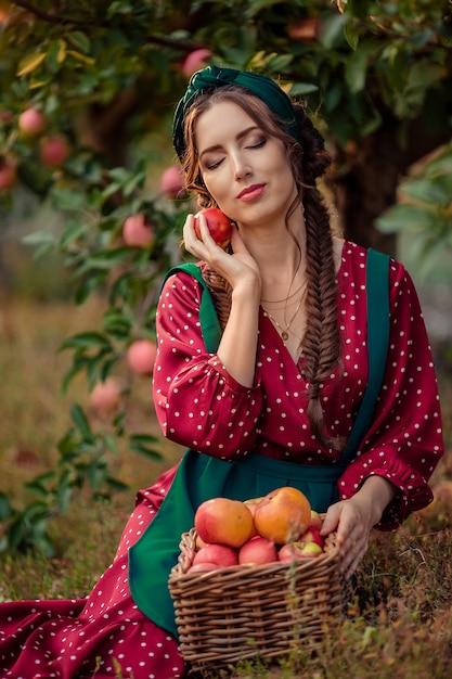 portrait of a woman in a red dress who sits near wicker baskets with ripe apples and holds one apple in her hands, closing her eyes. harvesting in the apple orchard