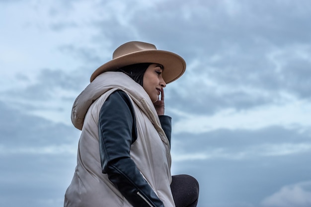 Portrait of a woman sideways holding her hat with her leg resting on a raised floor outdoors