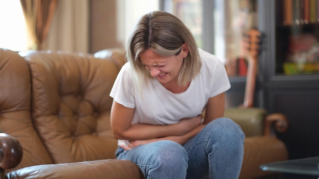 Portrait of woman sitting on sofa and touching belly writhing in pain stomachache menstrual