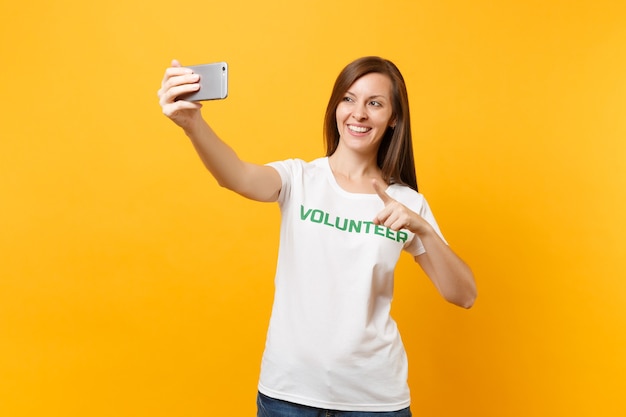 Portrait of woman in white t-shirt written inscription green title volunteer taking selfie shot on mobile phone isolated on yellow background. Voluntary free assistance help charity grace work concept