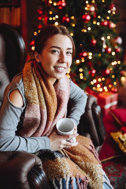 Portrait of Woman Who Sits in an Armchair Near Christmas Tree with Red Toys with Cup in Her Hands