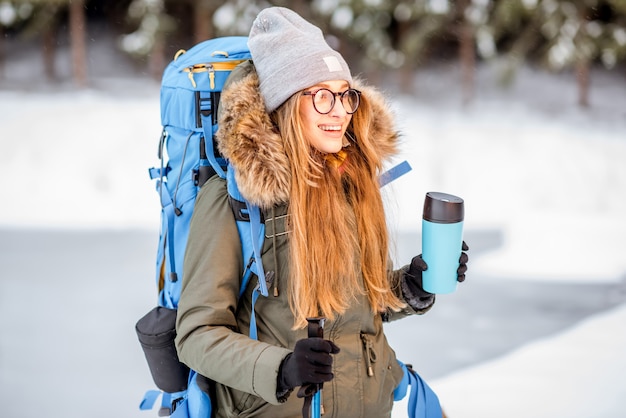 Photo portrait of a woman in winter clothes hiking with backpack, tracking sticks and thermos at the snowy forest near the frozen lake