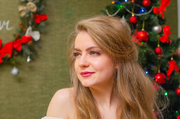 Portrait of a woman with bright red lips, blond long hair against the New Year tree. Young woman in a white warm coat, shoulders are visible. Holidays. Merry Christmas