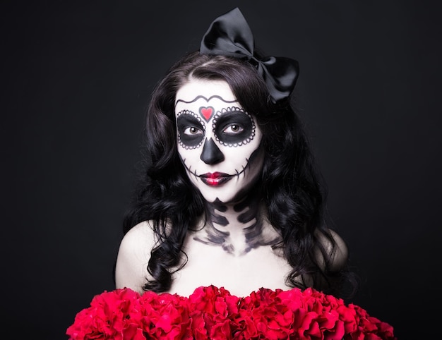 portrait of woman with creative Halloween skull make up and red flowers over black background