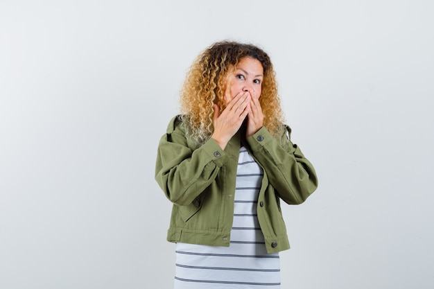 Portrait of woman with curly blonde hair keeping hands on mouth in green jacket and looking shocked front view