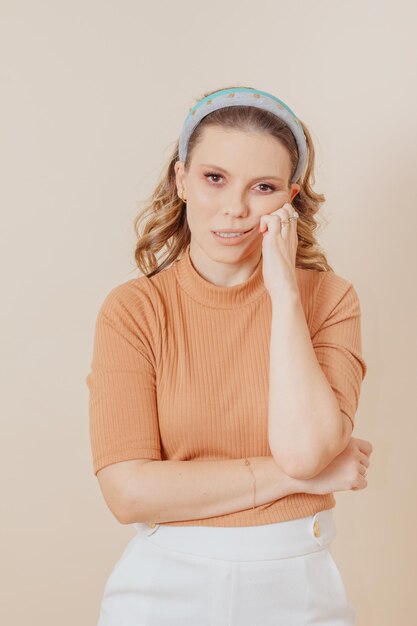 Portrait of woman with hand on chin with tired expression. Studio photo. Woman wears skin-colored blouse and white pants and two tiaras in her hair.