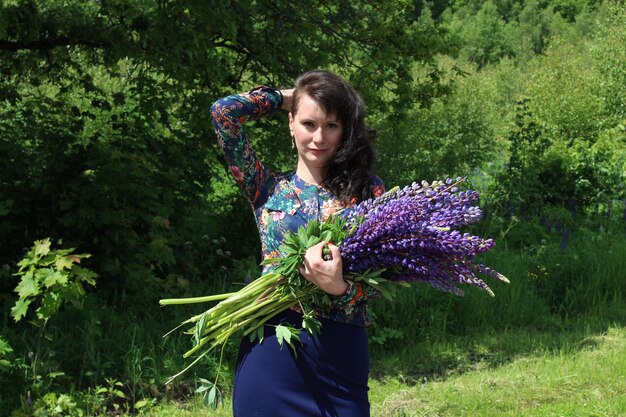 Photo portrait of woman with hand in hair holding lavenders while standing on field against trees