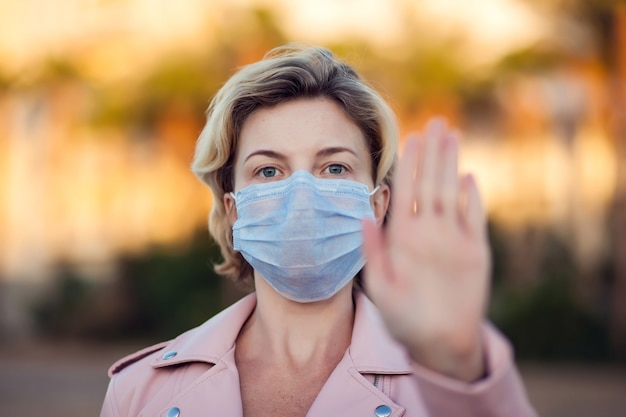 A portrait of woman with medical face mask showing stop sign with hands outdoor. People, healthcare and medicine concept