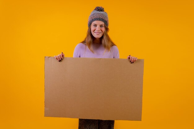 Portrait of a woman in a wool cap holding a cardboard sign on a yellow background