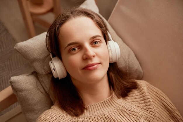 Portrait of young adult Caucasian beautiful woman with brown hair with relaxed face looking at camera listening to music with headphone while laying on sofa