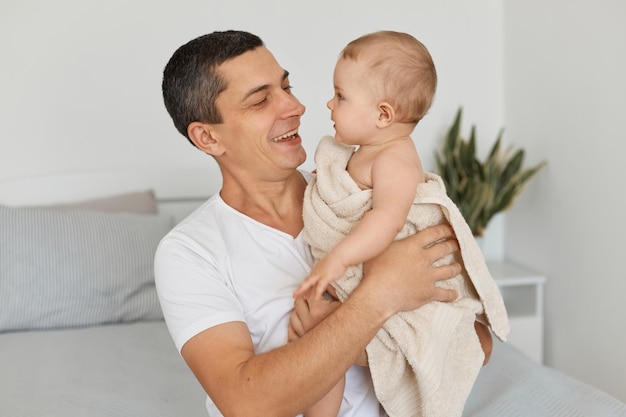 Portrait of young adult father with his toddler baby girl wrapped in towel, playing together at home, handsome man looking at his infant baby with toothy smile.