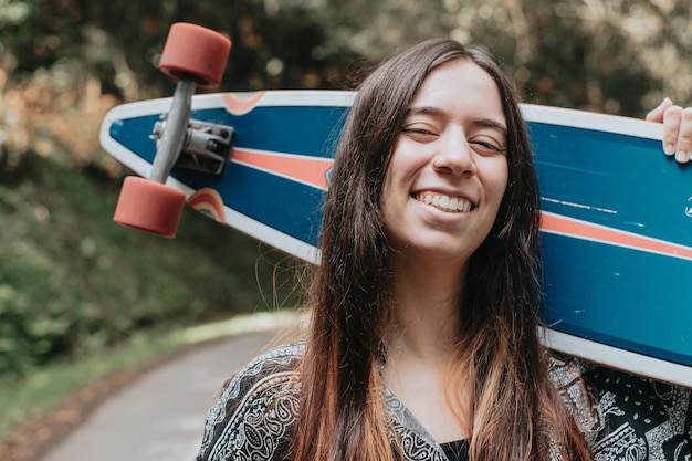 Portrait of young adult woman smiling with longboard skate outside on the road in the forestExcitedenthusiasticimpatient for new hobbyResting after a long travel doing sightseeingGeneration z