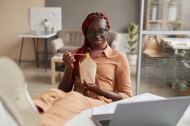 Portrait of young African-American woman eating takeout food and looking at laptop screen while relaxing at home office, copy space