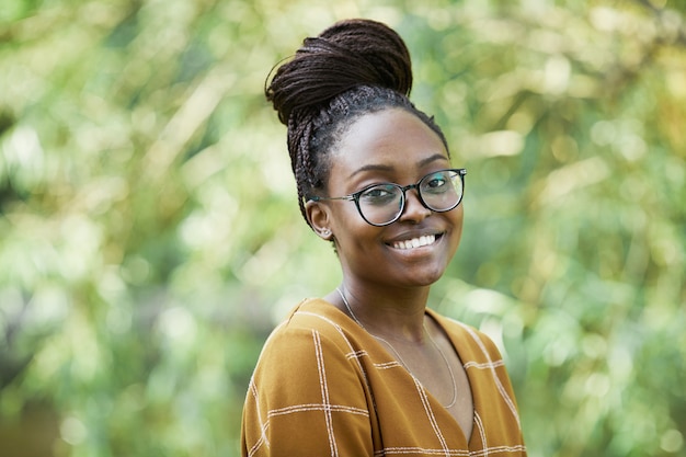 Portrait of young African-American woman wearing glasses and smiling at camera in park, copy space