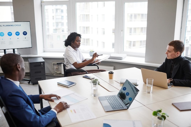 Portrait of young African-American woman in wheelchair leading business meeting in office