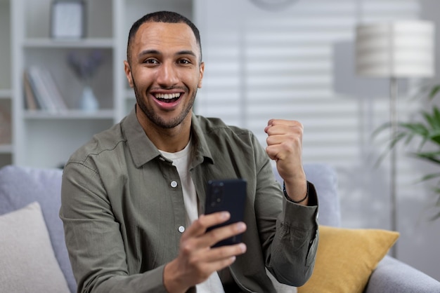 Portrait of a young africanamerican man using the phone sitting at home playing online games placing