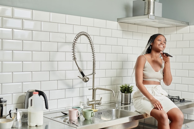 Portrait of young africanamerican woman speaking by smartphone while standing in kitchen interior co...