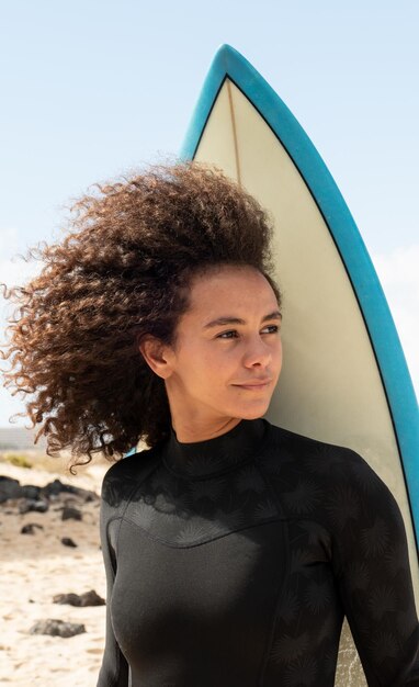 Portrait of a young afro surfer girl with surfboard on the beach wearing wetsuit