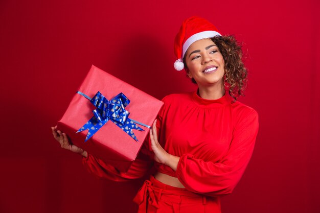 Portrait of young afro woman in santa claus hat holding a gift on red background. christmas night concept