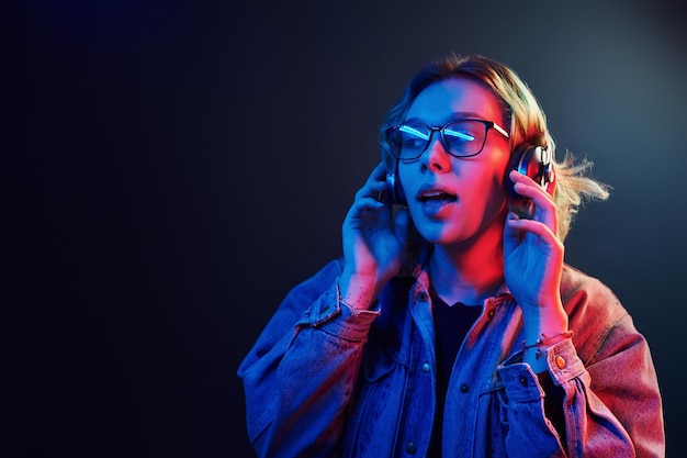 Portrait of young alternative girl in glasses with green hair in red and blue neon light in studio that listening to music in headphones