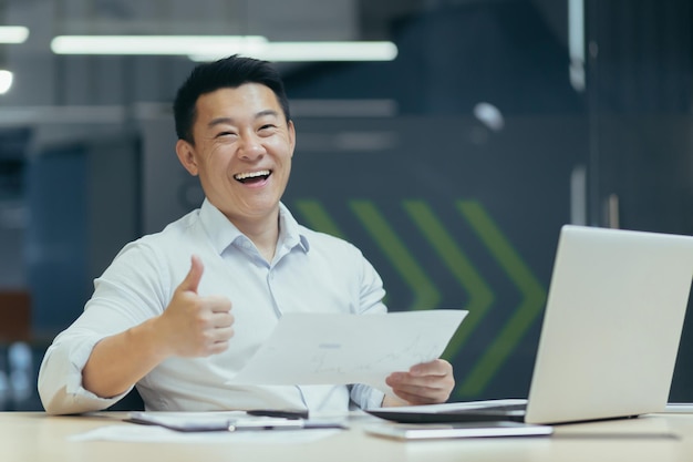 Portrait of a young Asian businessman man sitting at a desk in the office with a laptop and holding documents rejoices in a successful deal and increased income Pointing finger at camera super