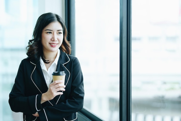 A portrait of a young Asian businesswoman smiling happily and drinking coffee by the window in the conference room