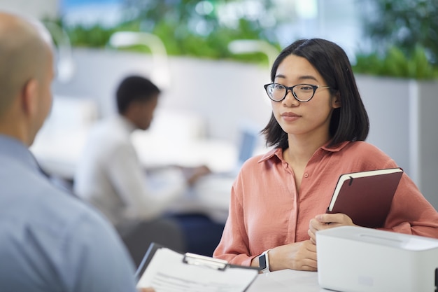 Portrait of young Asian businesswoman talking to boss or manager while standing in modern office