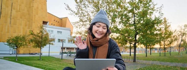 Portrait of young asian girl sits in warm clothes in park waves hand at tablet video chats outdoors