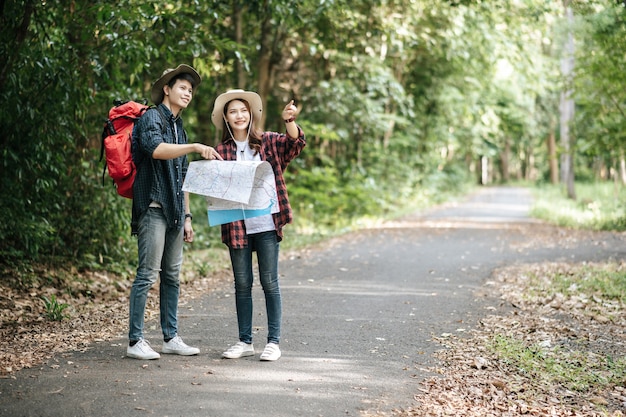 Portrait Young asian handsome man with backpack and trekking hat and pretty girlfriend standing and checking direction on paper map while walking on forest trail, backpack travel concept