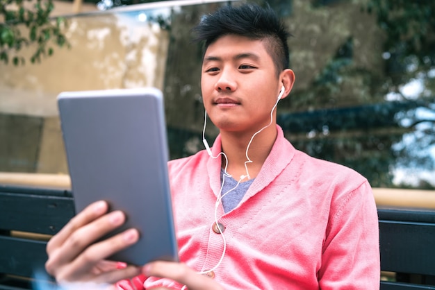 Portrait of young Asian man using his digital tablet with earphones while sitting in a bench outdoors. Technology concept.