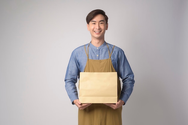 Portrait of young asian man wearing apron holding paper bag over white